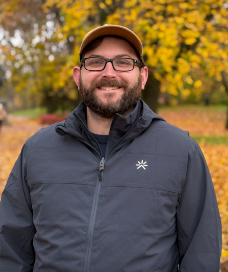 Ian Freng-Meeteer Headshot_glasses_edited Ian Freng-Meeteer owner of Occam's Razor Digital Marketing LLC wearing a rain coat and a hat in a fall setting with trees in the background.